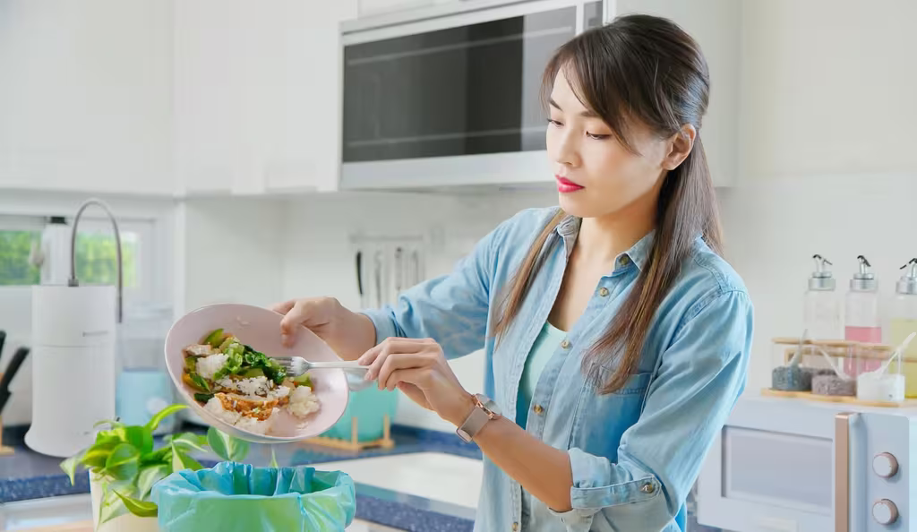 A woman throws leftover food into the trash, creating a food waste problem at home.