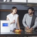 A joyful couple in the kitchen, with a woman ready to enjoy fruit while her husband prepares kiwi with the HomePure Viva Ioniser at the table